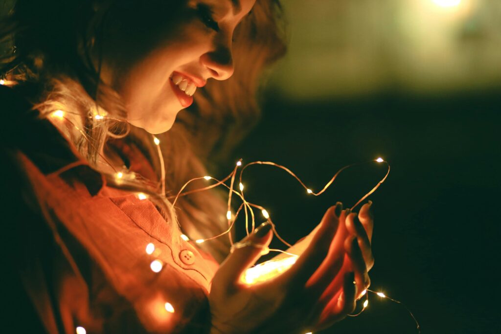 pexels photo 3450040 3450040 A smiling woman holds glowing fairy lights in a dark festive setting.